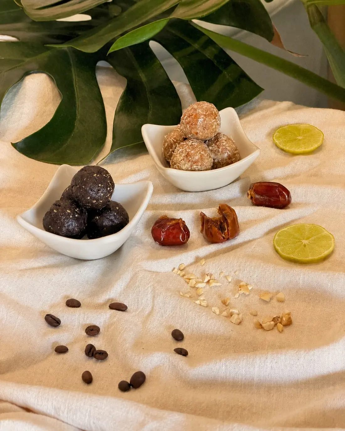 Dark cocoa and coconut bliss balls in white dishes with dates, lime and coffee beans on linen.