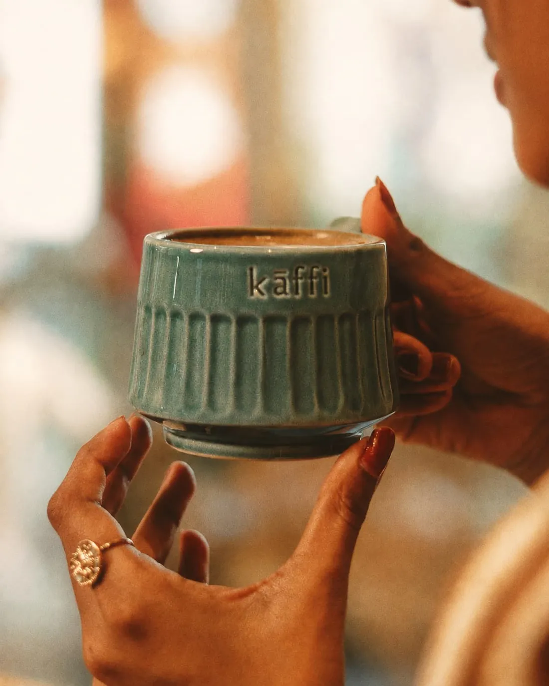 Two hands holding a teal glazed stoneware cup stamped kaffi.